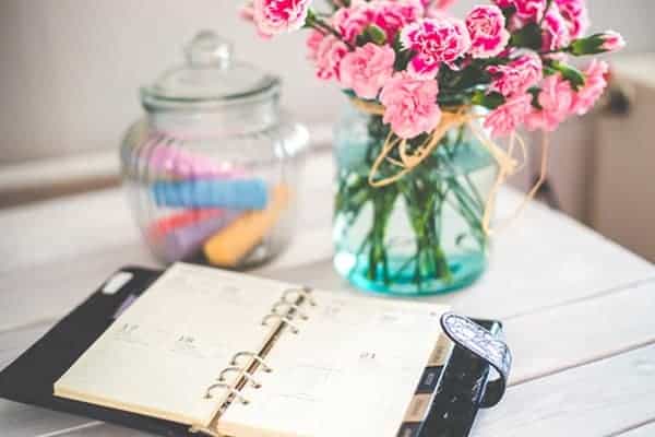 Freelancers black diary laid out on a desk next to a bunch of pretty pink flowers in a glass vase.