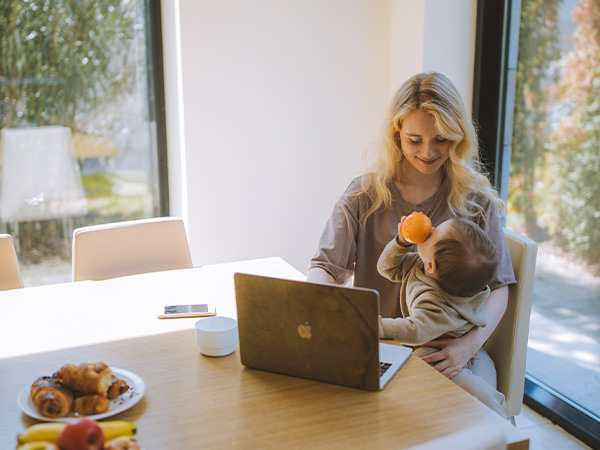 Mother working from home, with young baby sat on her knee and contently playing with an orange.