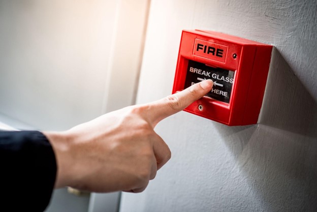 A man pointing at a red fire alarm switch on a concrete wall in an office building.